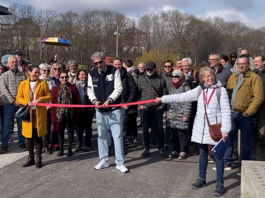 A group of smiling people while a man and woman hold and cut a red ribbon outside to mark the opening of a walkway or event. Trees and buildings can be seen in the background.