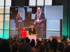 Zwei junge Frauen und ein Mann im Anzug stehen auf einer Bühne vor einem Publikum. Der Mann spricht von einem Podium aus, während auf einem großen Bildschirm hinter ihnen die gleiche Szene zu sehen ist.Fotos: Kerstin Ritscher, Silko Ritscher, Bodo