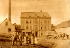 Ein Sepiafoto zeigt eine Gruppe von Menschen, darunter auch Kinder, die vor einem großen, dreistöckigen Gebäude stehen. In der Nähe stehen ein Pferdewagen und kleinere Gebäude, was auf eine historisch-ländliche Umgebung schließen lässt.