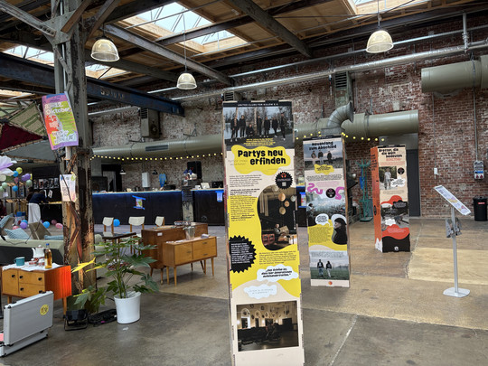 Ein überdachter Veranstaltungsraum mit freiliegenden Backsteinwänden und industriellen Balken bietet Informationstafeln, Retro-Möbel, Topfpflanzen und Menschen im Hintergrund. Das Sonnenlicht scheint durch die Oberlichter an der Decke.