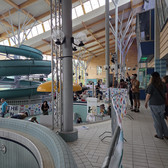 Die Menschen versammeln sich in einem modernen Hallenbad mit hohen Decken, natürlichem Licht und einer großen, grünen Wasserrutsche mit Spirale. Einige stehen an leeren Becken, andere sitzen an mit bunten Bannern geschmückten Tischen.