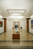 In a hallway with vaulted ceilings and tiled floors, there are works of art on the walls, a wooden bench with a book on it in the centre and a person walking away in the distance.