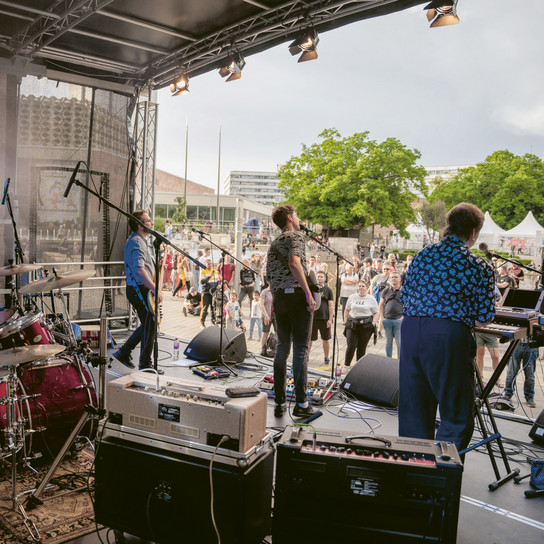 Eine Band mit Schlagzeug und Keyboards tritt auf einer Freilichtbühne auf. Die Musiker spielen für eine Menschenmenge, die sich auf einem Platz versammelt hat. Im Hintergrund sind Bäume und Gebäude zu sehen. Der Himmel ist bedeckt.