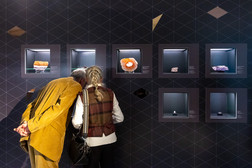 2 Personen stehen vor einer Wand mit Vitrinen. Darin sind große Minerale und Erze.