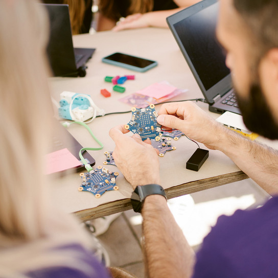 Zwei Personen arbeiten an einem Tisch mit Laptops und elektronischen Bauteilen in einem Klassenzimmer oder einer Werkstatt. Eine Person hält eine Platine, auf dem Tisch liegen Kabel und Haftnotizen.