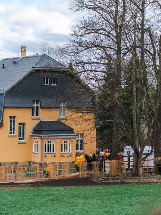 Foto: Kunstsammlungen Chemnitz/Ernesto Uhlmann/radar studios Ein großes gelbes Haus mit dunklem Dach ist von kahlen Bäumen und einer Wiese umgeben. Gelbe Luftballons schmücken den umzäunten Hof, und mehrere Menschen haben sich in der Nähe des Eingangs versammelt. Der Himmel ist teilweise bewölkt.