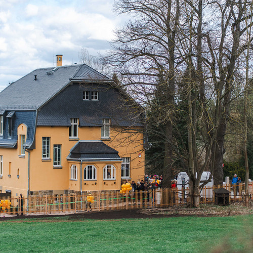 Foto: Kunstsammlungen Chemnitz/Ernesto Uhlmann/radar studios Ein großes gelbes Haus mit dunklem Dach ist von kahlen Bäumen und einer Wiese umgeben. Gelbe Luftballons schmücken den umzäunten Hof, und mehrere Menschen haben sich in der Nähe des Eingangs versammelt. Der Himmel ist teilweise bewölkt.