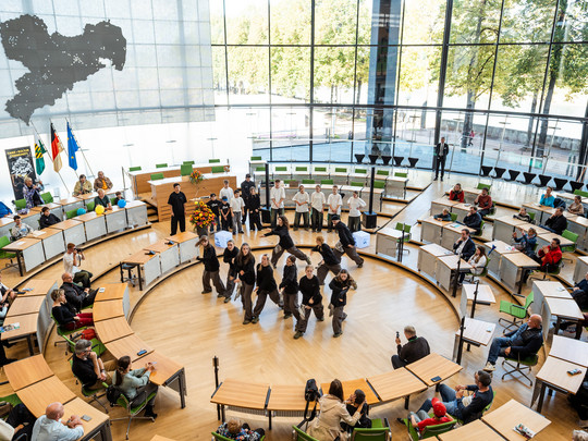 Eine Gruppe führt in der Mitte eines runden, modernen Auditoriums mit großen Fenstern einen Tanz auf. Die Leute sitzen herum und schauen zu, und auf der linken Seite sind Fahnen aufgestellt. Draußen sind Bäume und Sonnenlicht zu sehen.