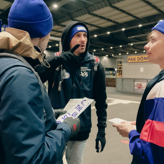 Drei Personen mit blauen Mützen und Jacken stehen an einem Bahnhof und unterhalten sich. Eine Person hält eine Broschüre, eine andere zeigt darauf und die dritte hört aufmerksam zu. Im Hintergrund sind ein Schild und ein Essbereich zu sehen.