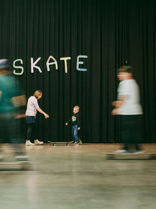 Ein Kind steht mit einem Erwachsenen vor einem schwarzen Vorhang, über dem das Wort "SKATE" steht. Mehrere unscharfe Personen auf Skateboards bewegen sich um sie herum auf einem glatten Hallenboden.