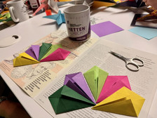 Bunte Origami-Papierfächer liegen auf einem aufgeschlagenen Buch neben einer Schere, während im Hintergrund eine Tasse und weitere Origami-Papiere und Kreationen auf dem Tisch verstreut sind.