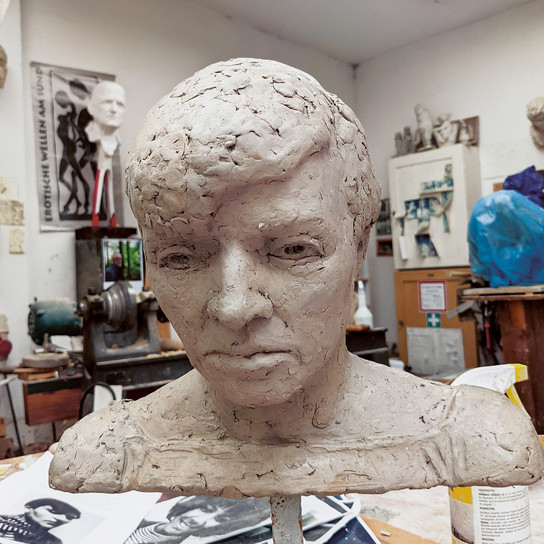 Eine Tonbüste eines menschlichen Kopfes mit detaillierten Gesichtszügen und strukturiertem Haar steht auf einem Tisch in einem Kunstatelier. Im Hintergrund sind verschiedene Werkzeuge, Kunstwerke und Skulpturen zu sehen.