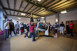 Eine Gruppe von Menschen in legerer Kleidung übt in einem geräumigen, gut beleuchteten Raum mit Holzfußboden und -decke Akro-Yoga oder Gruppenakrobatik, wobei einige eine menschliche Pyramide bilden und andere zusehen oder warten, bis sie an der Reihe sind.