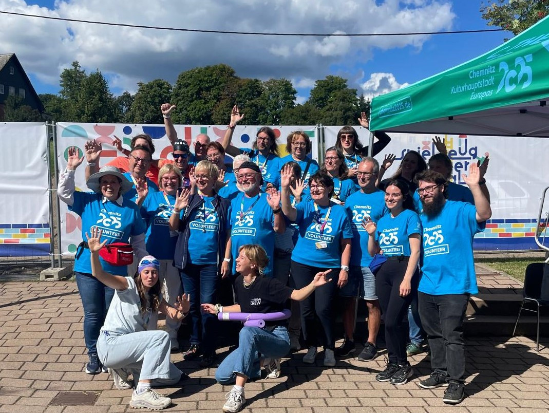 Eine Gruppe von Menschen in passenden blauen Hemden posiert und winkt unter einem sonnigen Himmel bei einer Veranstaltung im Freien. Sie stehen in der Nähe eines Zeltes mit der Aufschrift "25" und bunten Dekorationen im Hintergrund.