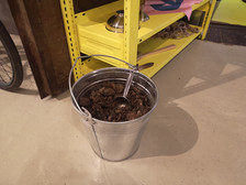 Ein Metalleimer, gefüllt mit brauner Erde oder Kompost und einer Metallschaufel darin, steht auf einem beigen Boden vor einem gelben Regal.