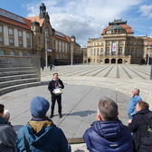 A tour guide speaks to a small group of people in an open, sunny town square, surrounded by historic buildings with ornate facades and red roofs. The sky is partly cloudy.
