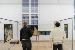 Zwei Personen stehen vor eingezäunten Kunstauslagen und betrachten Gemälde in einem hellen, modernen Galerieraum mit weißen Wänden, hohen Fenstern und Kunstwerken, die Menschen in gedeckten Farben darstellen.