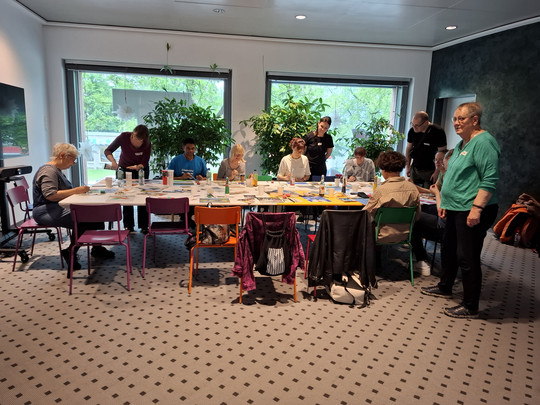 A group of people sit and stand in a bright room with large windows, plants and colourful chairs around a table covered with art supplies, engaged in a creative activity or workshop.