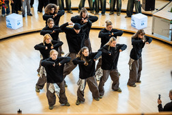 A group of dancers in wide brown trousers and black shirts perform a synchronised number on a wooden floor, while an audience and advertising displays can be seen in the background.