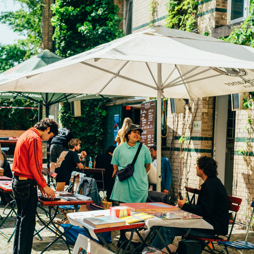 Menschen versammeln sich um Tische im Freien unter großen Sonnenschirmen in einem Kopfsteinpflasterbereich. Auf und um die Tische herum sind Kunstwerke und Gegenstände ausgestellt. Die Szene scheint ein zwangloses Markt- oder Café-Ambiente im Freien zu sein, mit einem Backsteingebäude und Grünflächen im Hintergrund.