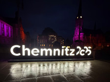 Large illuminated "Chemnitz 2025" sign at night on a town square, with historic buildings and a church in the background, both illuminated with purple lights. The wet pavement reflects the glow of the sign.
