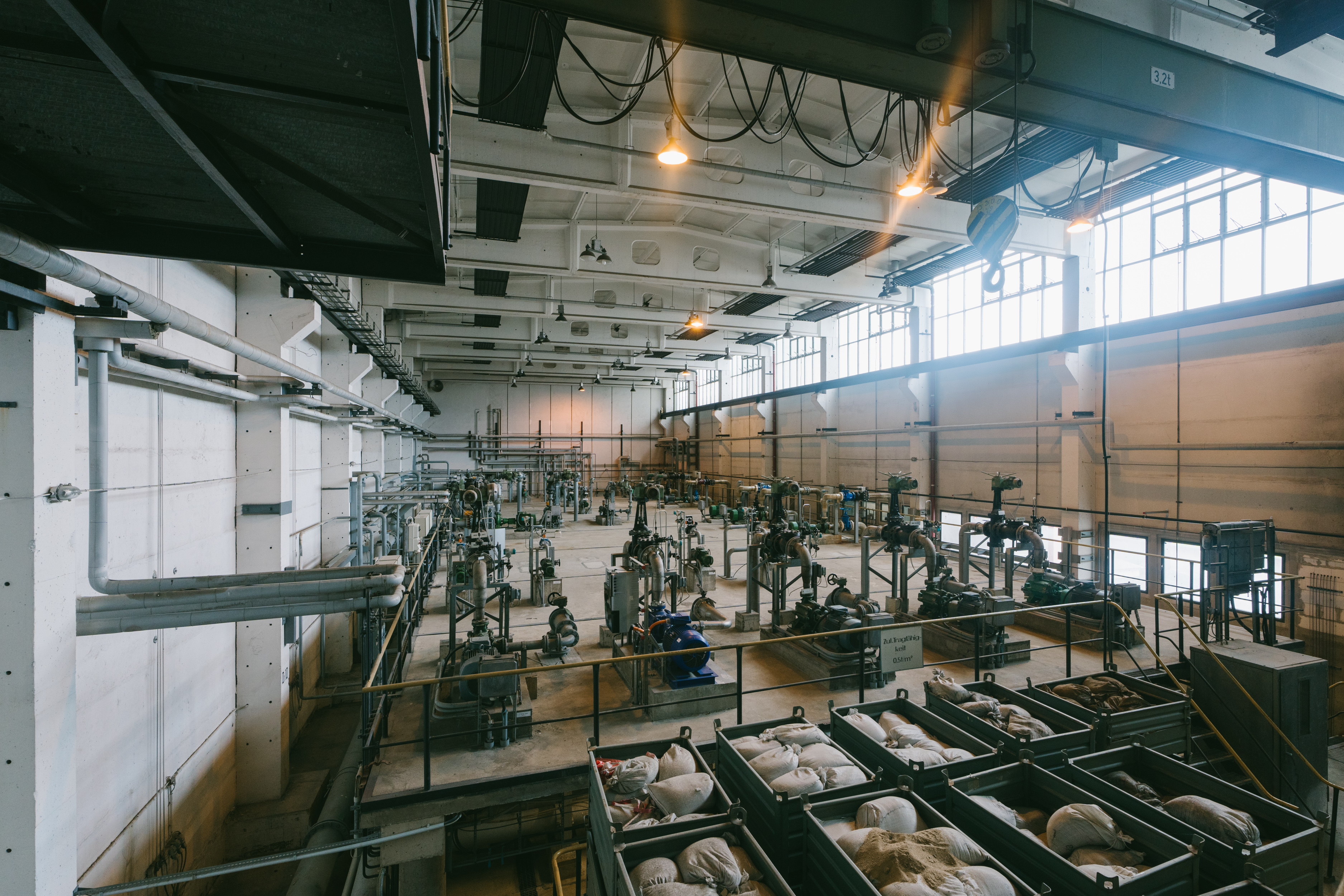 An extensive industrial plant with high ceilings, large ceiling lights and numerous large pipes and machines. Several large containers with white sacks can be seen in the foreground. Sunlight enters through high windows on the right-hand side.