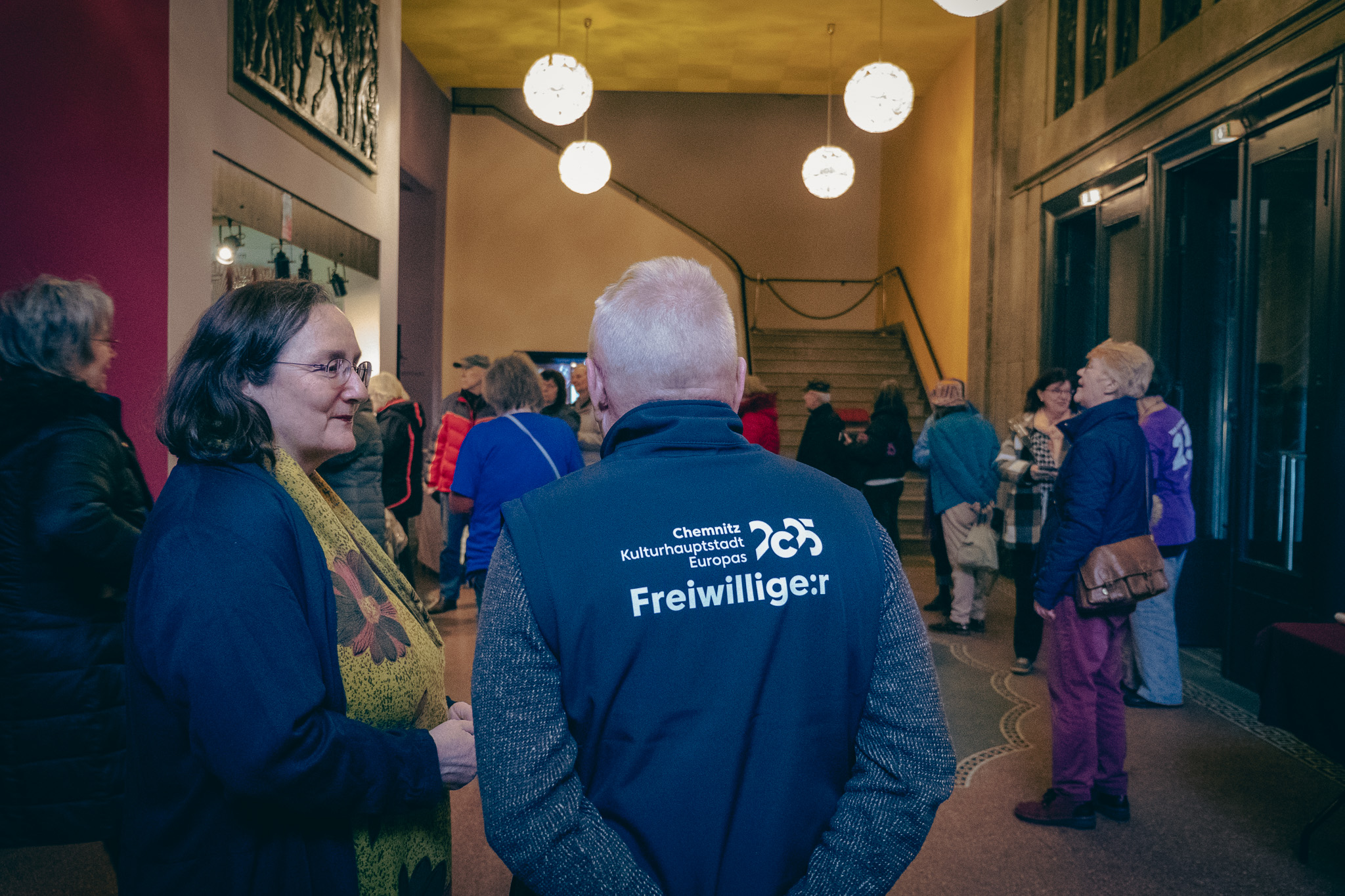 A group of people are standing in the entrance hall of a building and chatting. One person is wearing a waistcoat labelled "Volunteer". The atmosphere is relaxed; others are gathered or walking towards a staircase in the background. Spherical lights hang from the ceiling.
