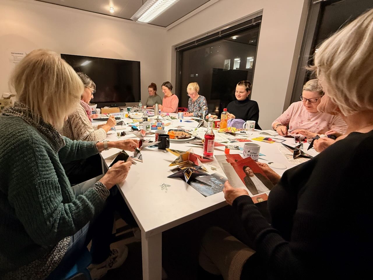 A group of people are sitting in a well-lit room around a table covered with craft materials, magazines and scissors, doing handicrafts.