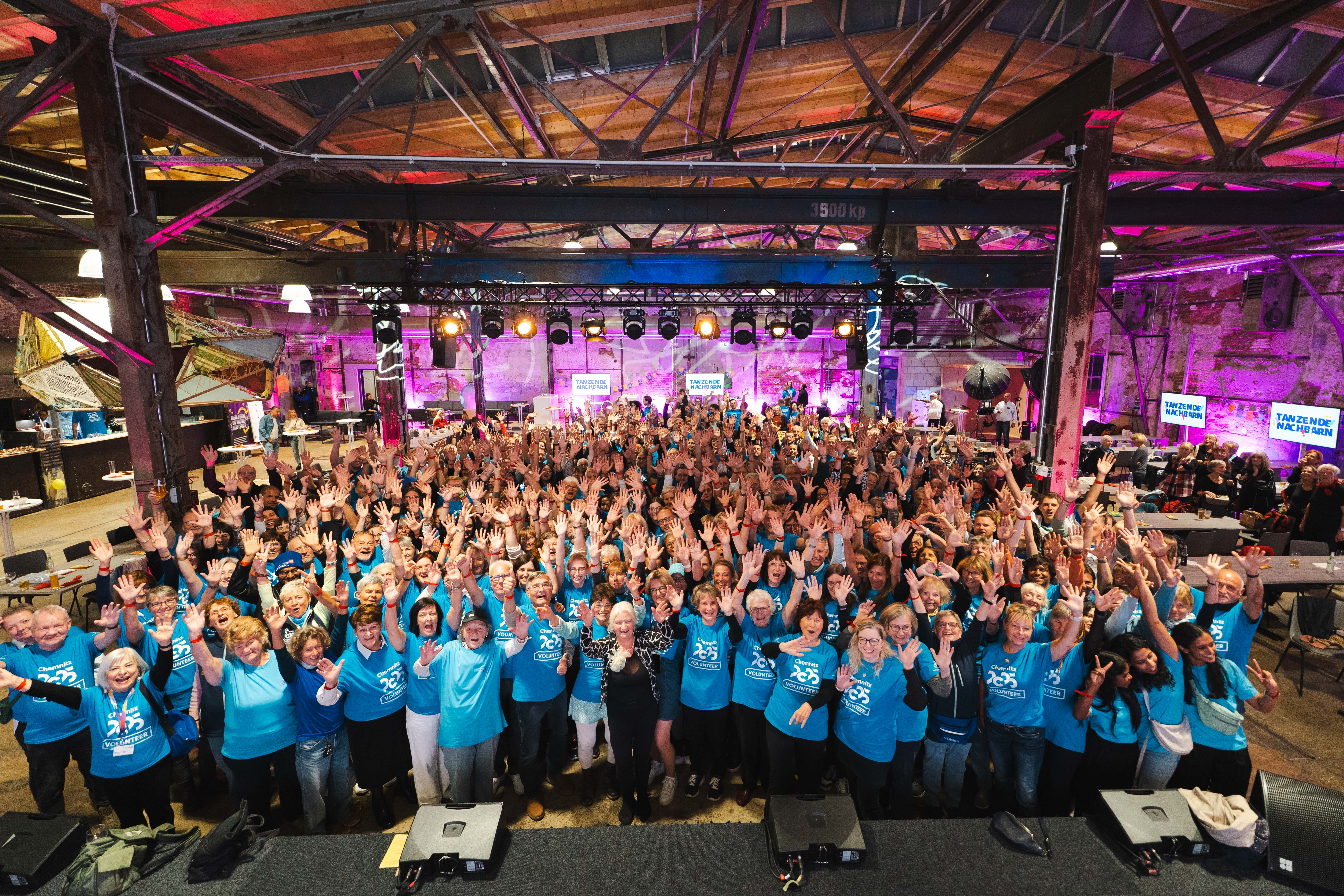 Eine große Gruppe von Menschen in passenden blauen Hemden steht in einem Innenraum, lächelt und winkt in die Kamera bei einer Veranstaltung unter einer Decke im industriellen Stil mit hellen Lichtern und einer Bühne im Hintergrund.