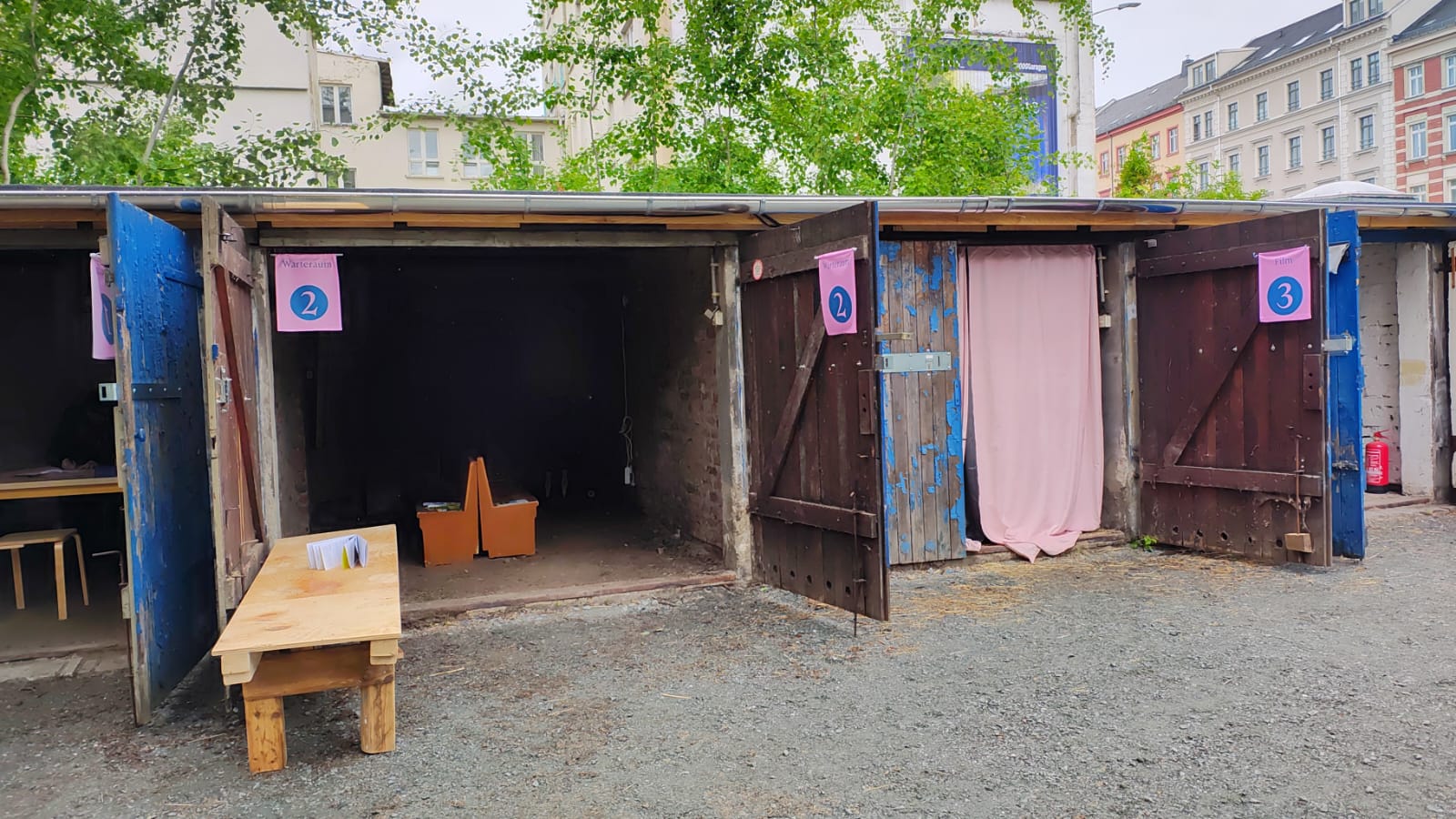 Three wooden stands with open doors are labelled with the numbers 1, 2 and 3. Stand 1 has a table with papers in front of it, stand 2 is empty and stand 3 has a pink curtain. The area is covered with gravel flooring and buildings in the background.