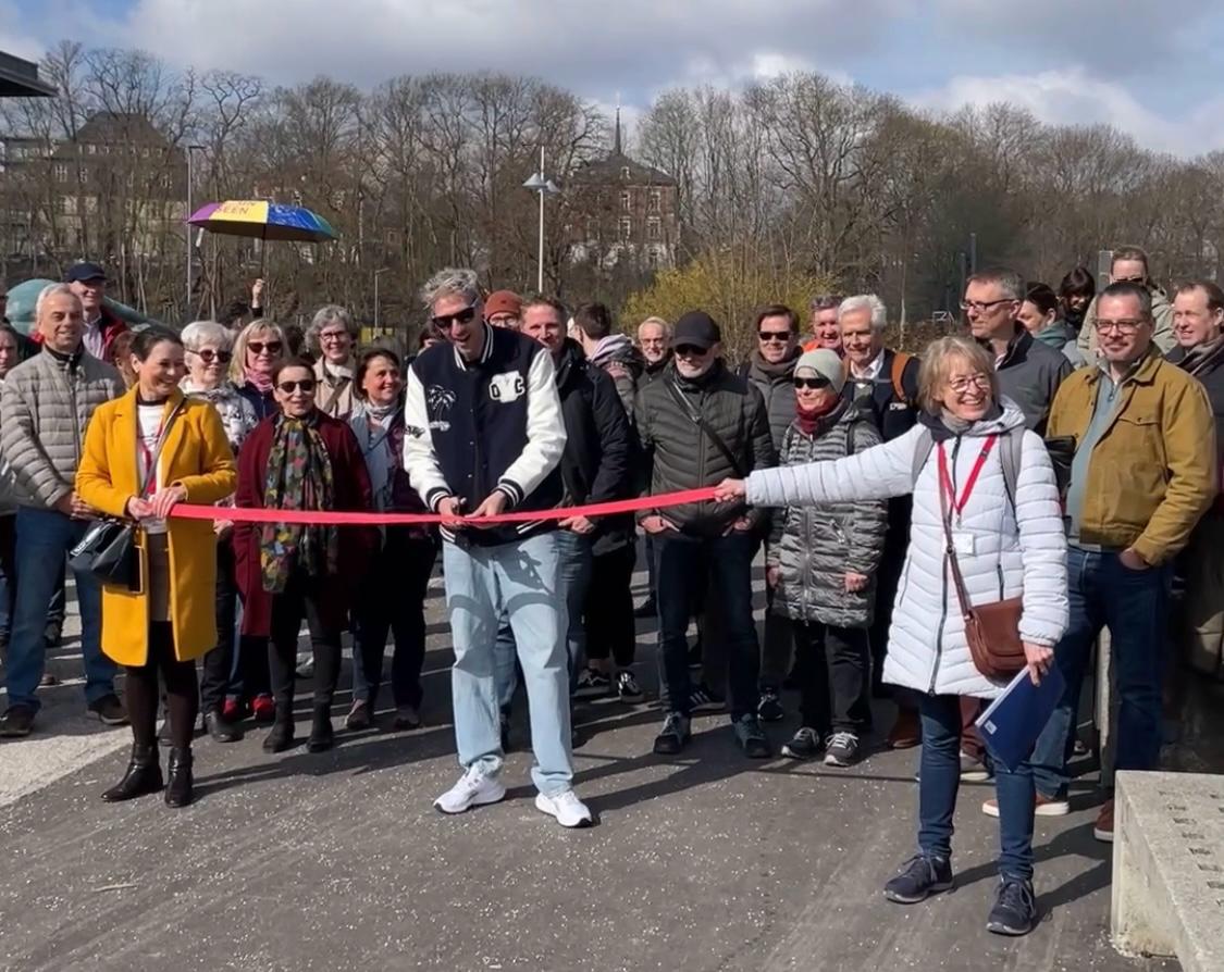 A group of smiling people while a man and woman hold and cut a red ribbon outside to mark the opening of a walkway or event. Trees and buildings can be seen in the background.